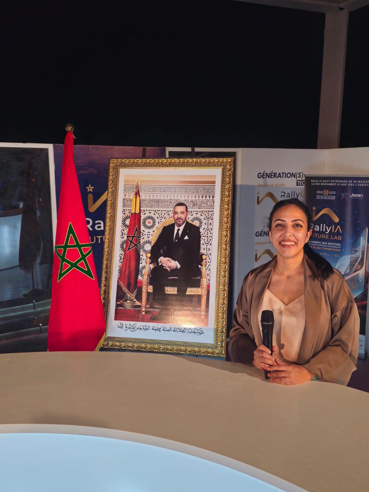 Le Dr Sana Amraoui, micro en main et souriante, sur le plateau du Rally IA Future Lab à Merzouga, devant le drapeau marocain et le portrait de Sa Majesté le Roi Mohammed VI